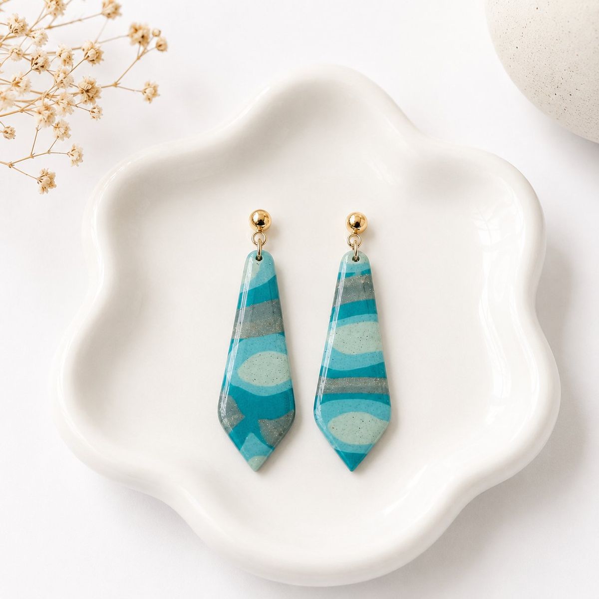 Two elongated pointed clay dangle earrings with teal, mint, and grey swirl patterns hanging from gold ball studs, displayed on a white ceramic dish.