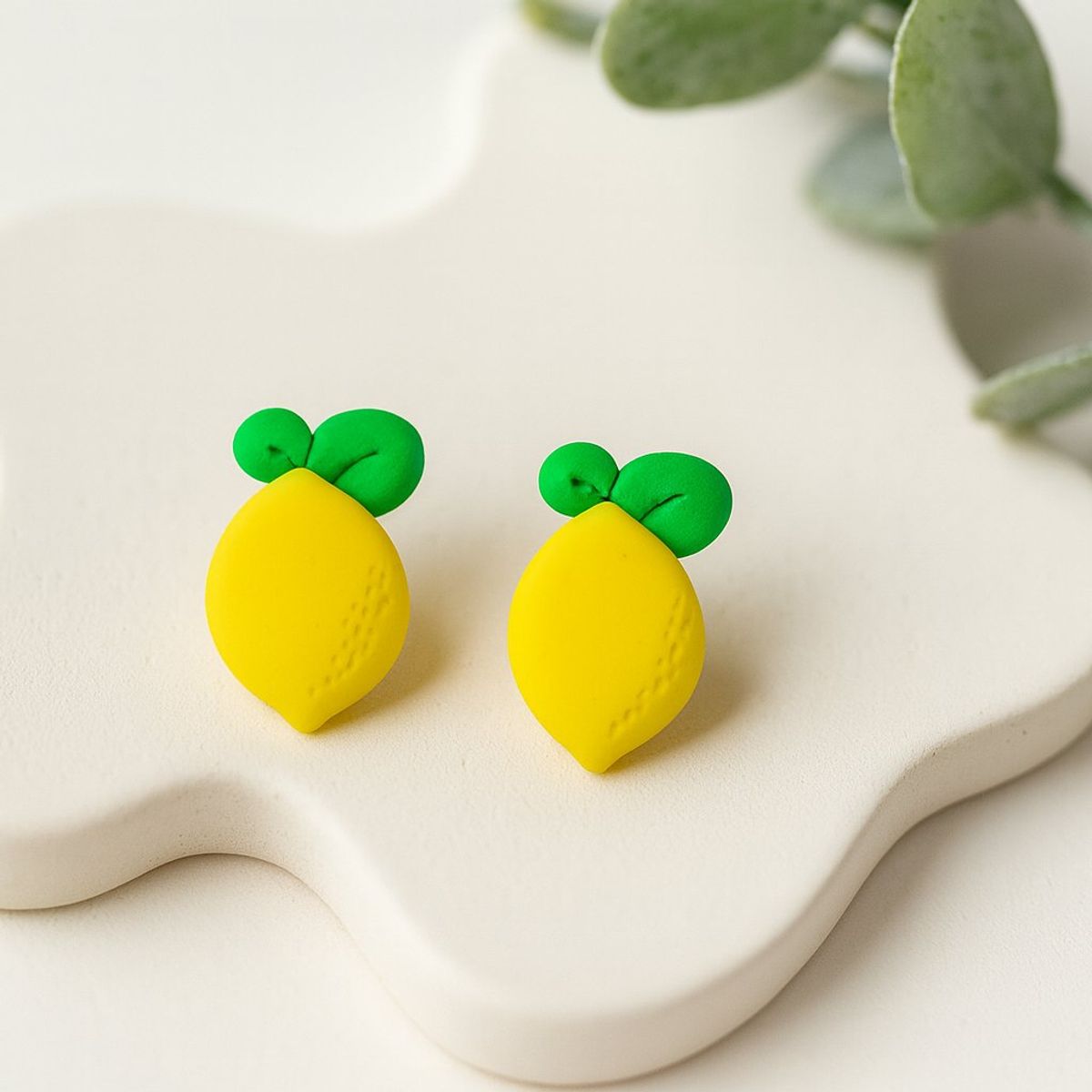 A pair of bright yellow lemon-shaped polymer clay stud earrings with green leaf details displayed on a white ceramic tray.