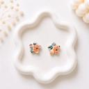 A pair of white clay flower-shaped stud earrings with an orange floral and green leaf print, displayed on a white wavy ceramic tray.
