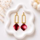 A pair of dangle earrings with gold arch connectors and ball studs, holding diamond-shaped clay pendants in deep red with pink swirl patterns, displayed on a white ceramic tray.