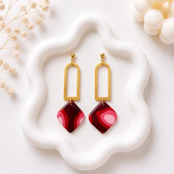 A pair of dangle earrings with gold arch connectors and ball studs, holding diamond-shaped clay pendants in deep red with pink swirl patterns, displayed on a white ceramic tray.