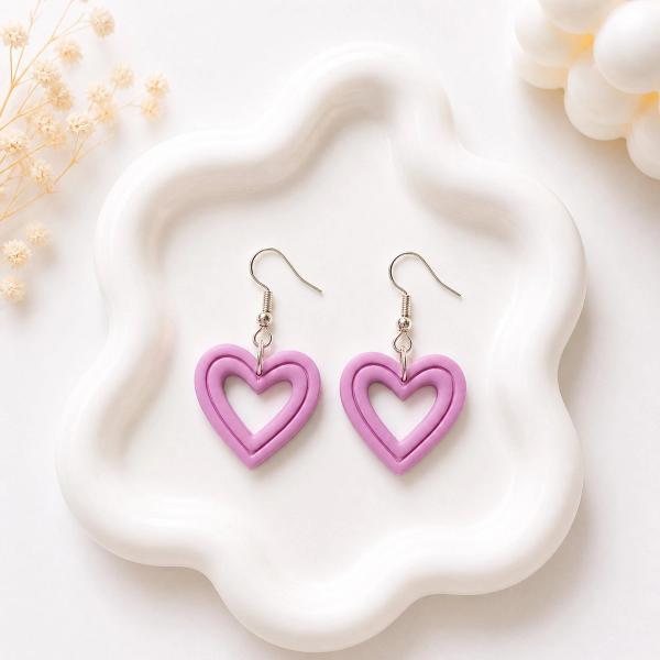 A pair of lavender clay open-heart dangle earrings with silver hook findings displayed on a white wavy tray.