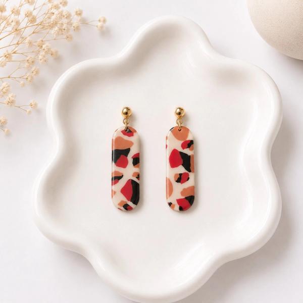 A pair of elongated pill-shaped clay dangle earrings with a red, black, and terracotta terrazzo pattern on a cream base, hanging from gold ball stud posts, displayed on a white ceramic dish.