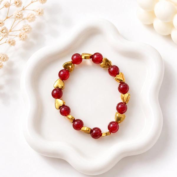 A stretch bracelet with round red translucent beads and gold-toned heart spacers displayed on a white ceramic dish.