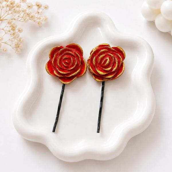 Two handmade clay rose bobby pins with red petals and gold edges displayed on a white ceramic tray.