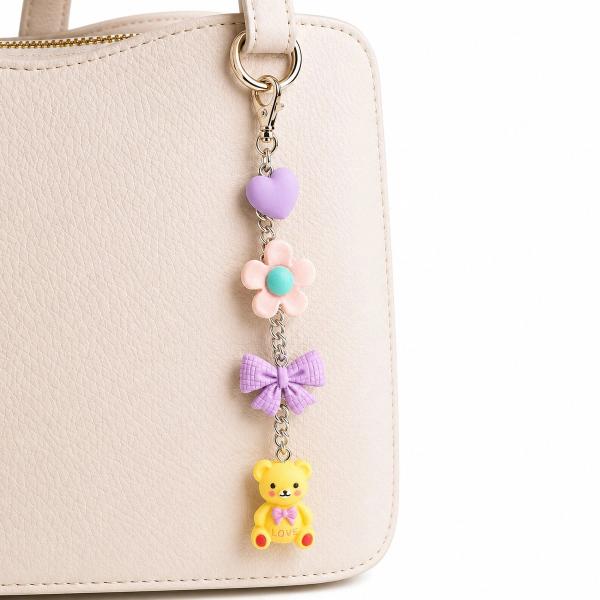 A pastel bag charm hanging from a cream handbag strap, featuring a lavender heart, pink daisy, purple bow, and yellow teddy bear charms connected by a silver chain.