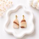 A pair of trapezoid-shaped clay dangle earrings with cream and brown marbled stripes hanging from gold ball studs, displayed on a white wavy ceramic tray.