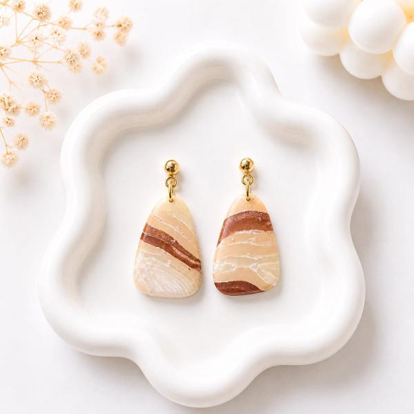 A pair of trapezoid-shaped clay dangle earrings with cream and brown marbled stripes hanging from gold ball studs, displayed on a white wavy ceramic tray.