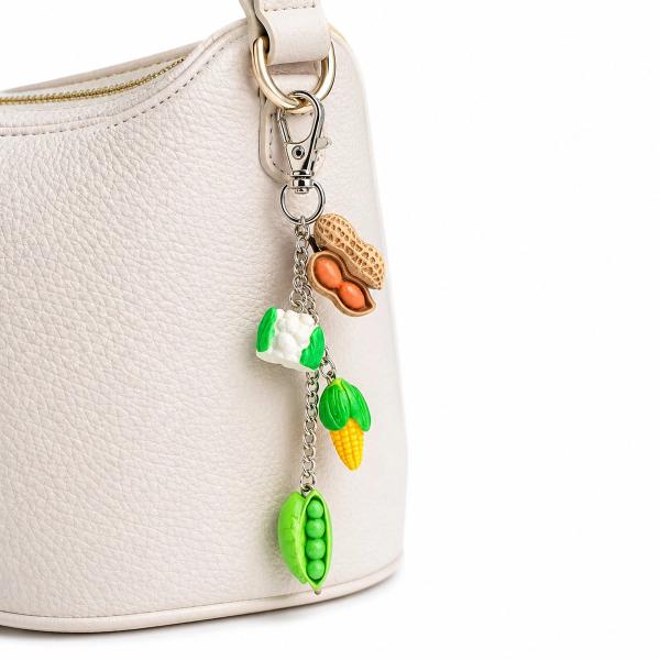 A polymer clay bag charm with four miniature vegetable charms — peanut, cauliflower, corn, and pea pod — on a silver chain, clipped to a cream leather handbag.
