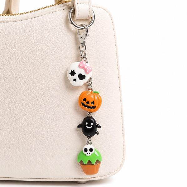 A silver-clasp bag charm with four Halloween polymer clay charms — a white skull with pink bow, orange jack-o'-lantern, black ghost, and a skull cupcake — clipped onto a cream handbag.