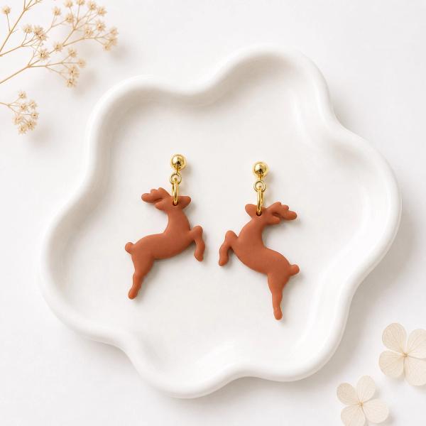 A pair of terracotta brown clay reindeer dangle earrings with gold ball studs and jump rings, displayed on a white scalloped dish.