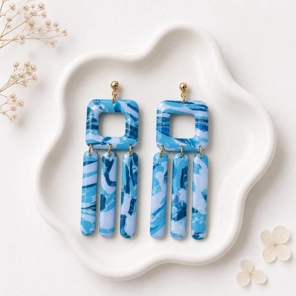 A pair of blue and white marbled clay dangle earrings with an open square top and three bar pendants hanging from gold jump rings on a white ceramic dish.