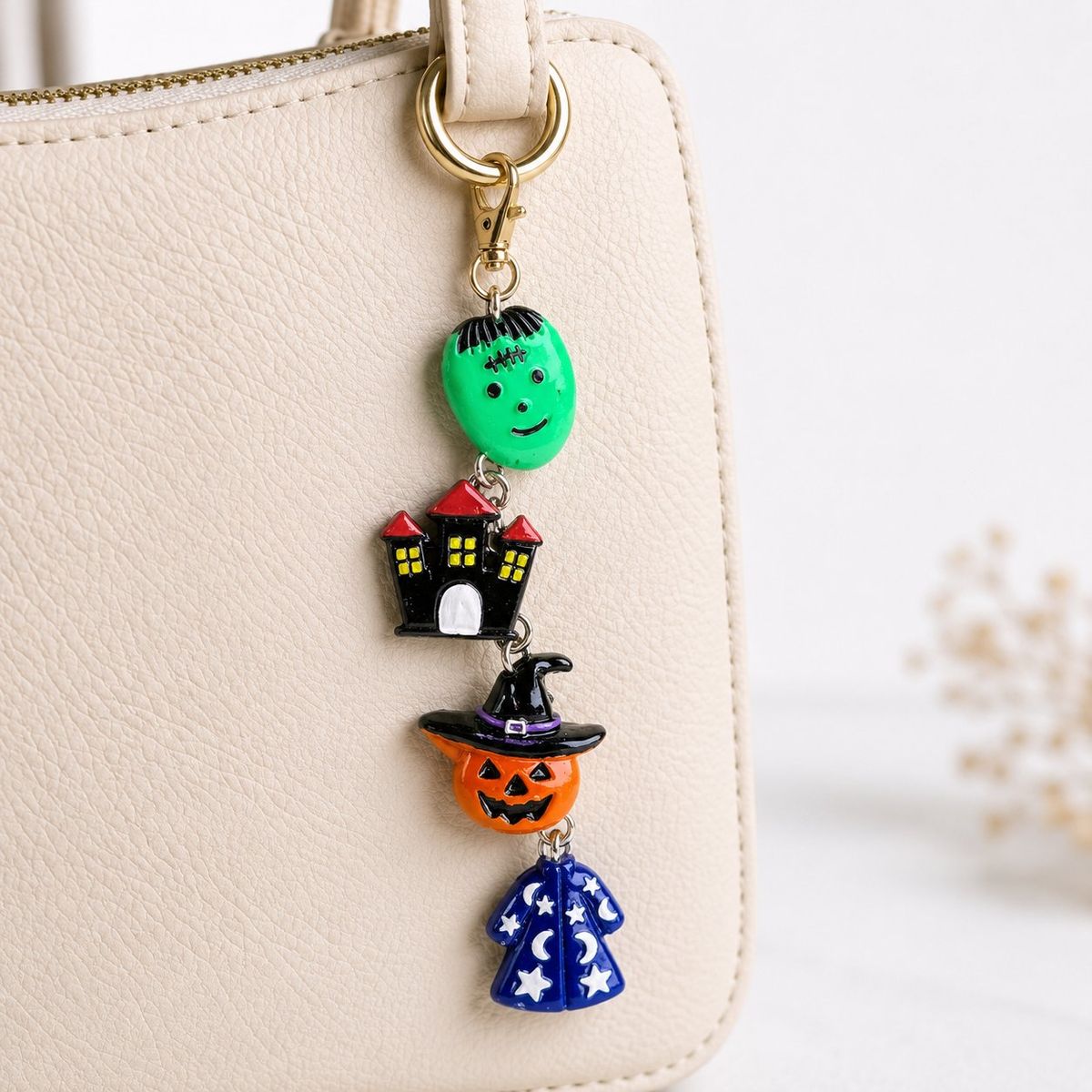 A stacked Halloween key charm featuring a green Frankenstein face, black haunted house, orange jack-o'-lantern with witch hat, and blue wizard cloak, clipped to a cream handbag with a gold lobster clasp.