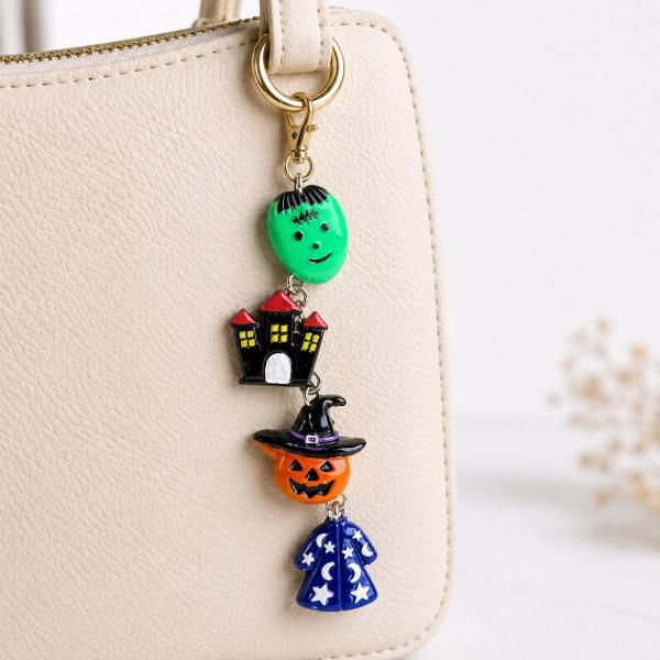 A stacked Halloween key charm featuring a green Frankenstein face, black haunted house, orange jack-o'-lantern with witch hat, and blue wizard cloak, clipped to a cream handbag with a gold lobster clasp.