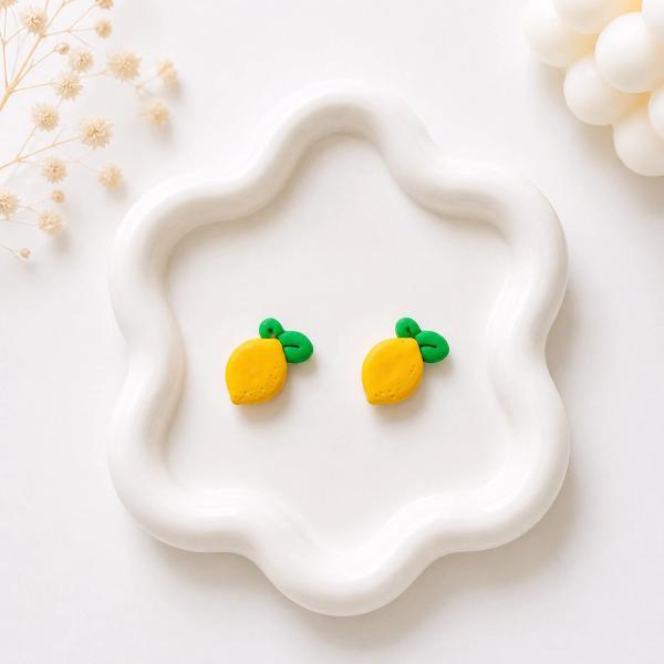 A pair of small yellow lemon-shaped polymer clay stud earrings with green leaf accents displayed on a white wavy ceramic tray.