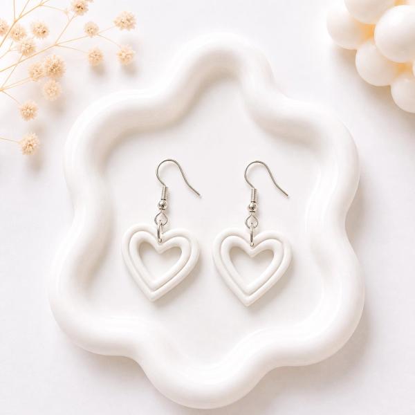 A pair of white clay open-heart dangle earrings with silver fish hook closures displayed on a white wavy tray.