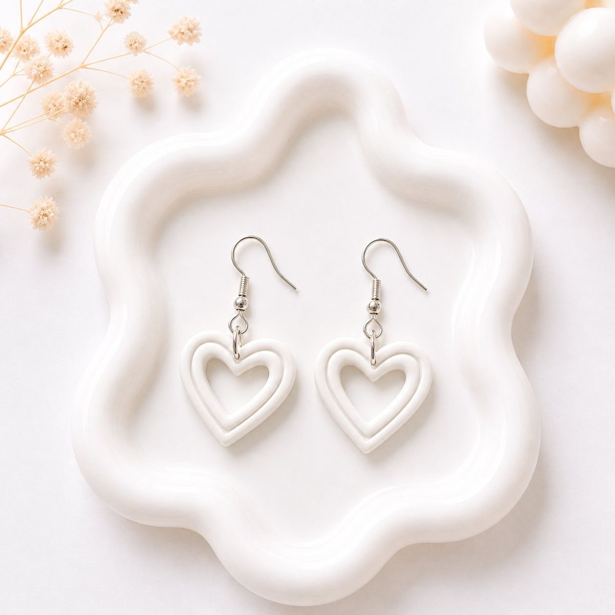 A pair of white clay open-heart dangle earrings with silver fish hook closures displayed on a white wavy tray.