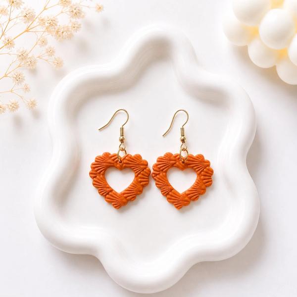A pair of burnt orange polymer clay open-heart dangle earrings with a ridged scalloped texture and gold hook findings, displayed on a white ceramic tray.
