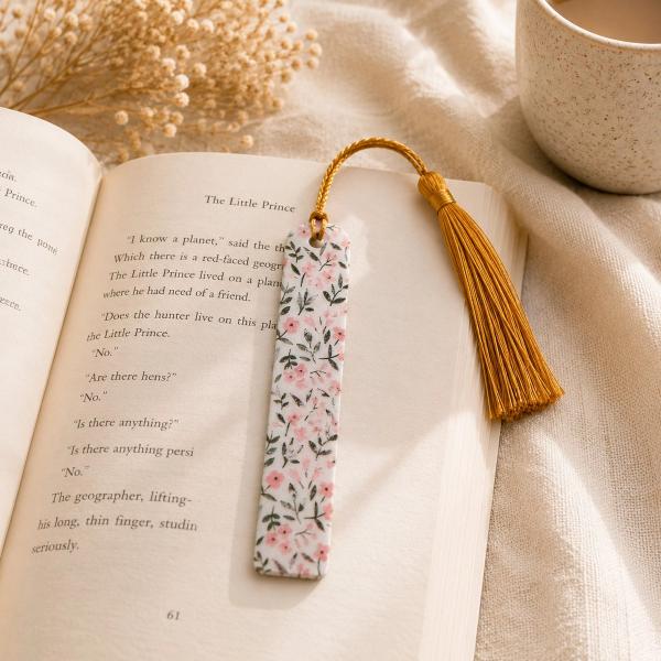 A white polymer clay bookmark with a pink floral and green leaf pattern, attached to a gold tassel, resting on an open book.