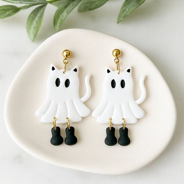 A pair of white polymer clay ghost-cat dangle earrings with black eyes and small black boot charms hanging from gold hoops, displayed on a white ceramic dish.