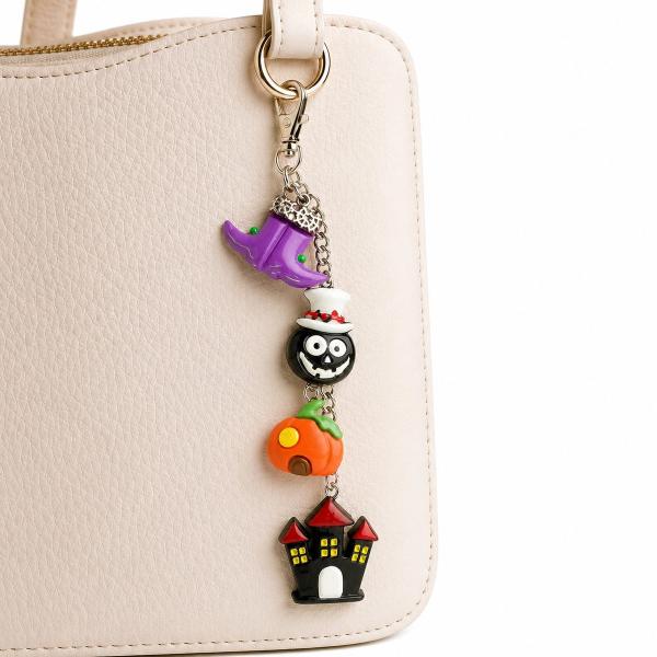 A dangling Halloween bag charm with four polymer clay charms — a purple witch hat, black skull with white top hat, orange pumpkin, and black haunted house — attached to a cream leather bag via a silver lobster clasp.