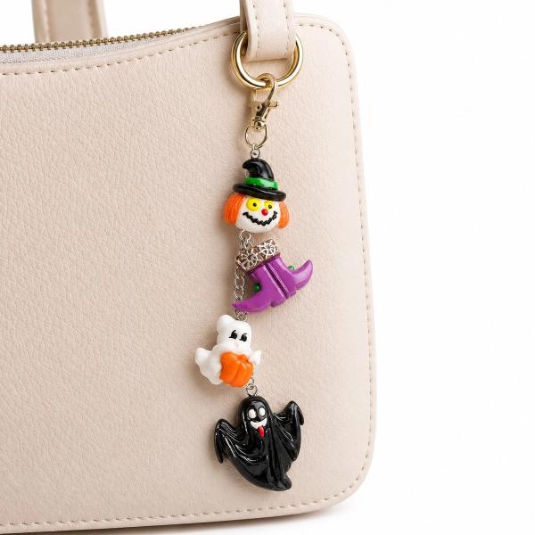 A Halloween bag charm with four resin charms — a witch head, purple witch boots, a white ghost with pumpkin, and a black ghost — hanging from a gold lobster clasp on a cream handbag.