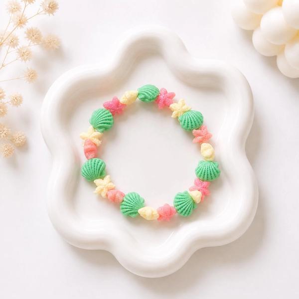 A circular stretch bracelet made of green scallop shell, pink starfish, and cream coral-shaped beads displayed on a white flower-shaped ceramic tray.