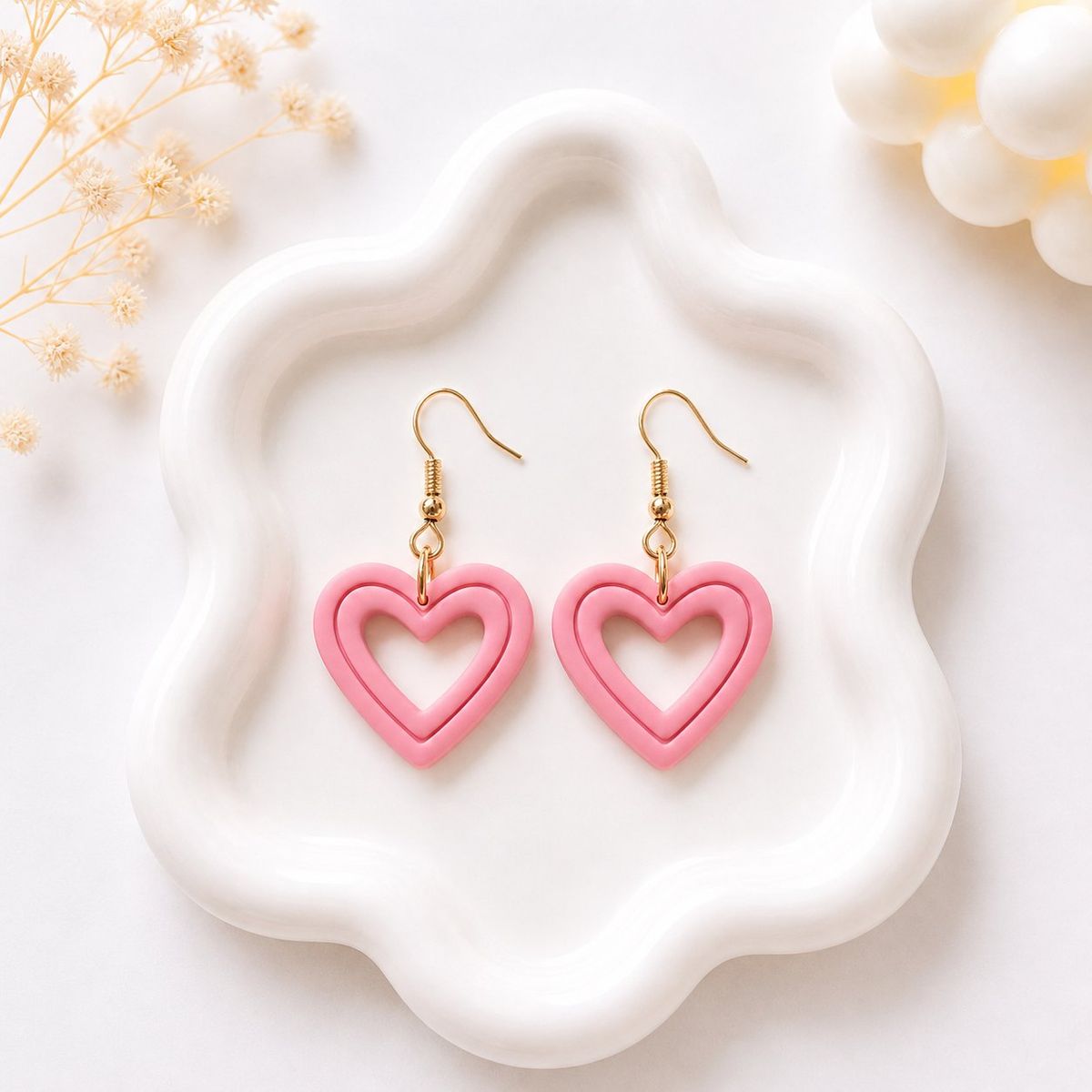 A pair of pink clay open-heart dangle earrings with gold fish hook wires displayed on a white wavy ceramic tray.