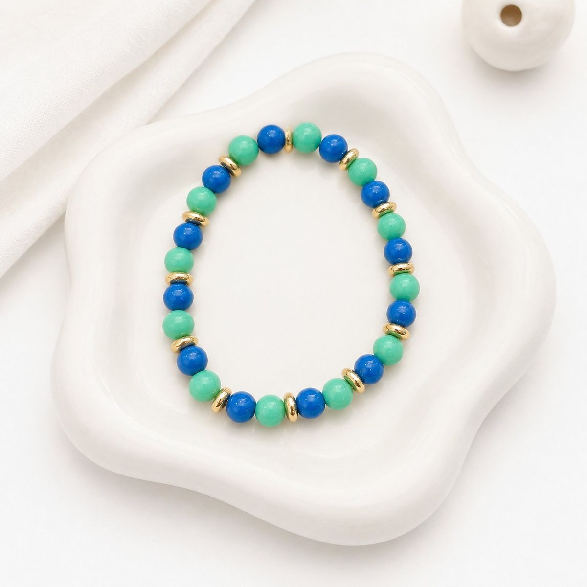 A stretch bracelet with alternating cobalt blue and mint green round beads separated by small gold disc spacers, laid flat on a white ceramic tray.