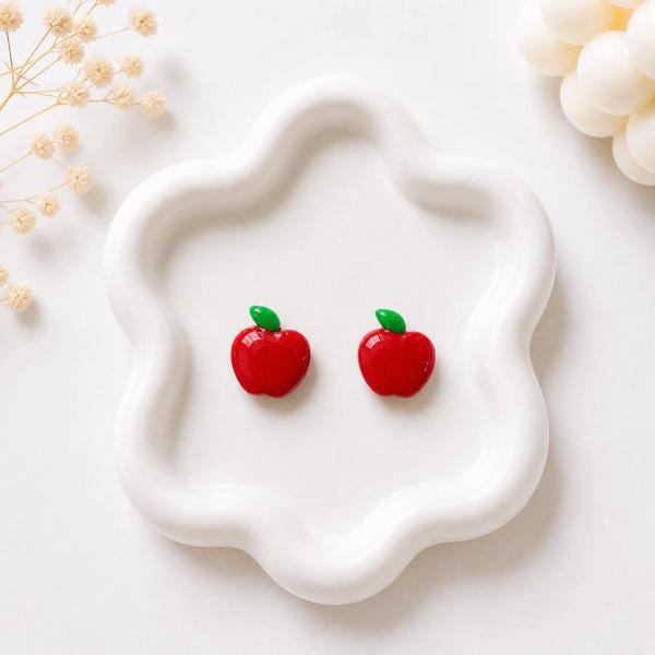 A pair of small red apple-shaped stud earrings with green leaf details displayed on a white ceramic tray.