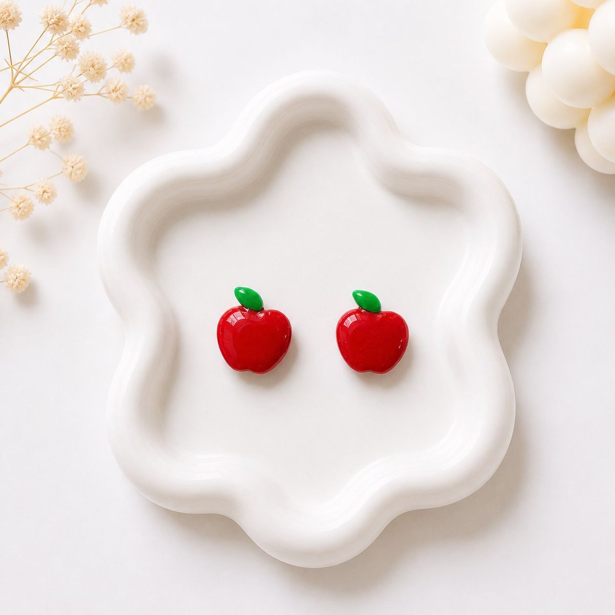 A pair of small red apple-shaped stud earrings with green leaf details displayed on a white ceramic tray.