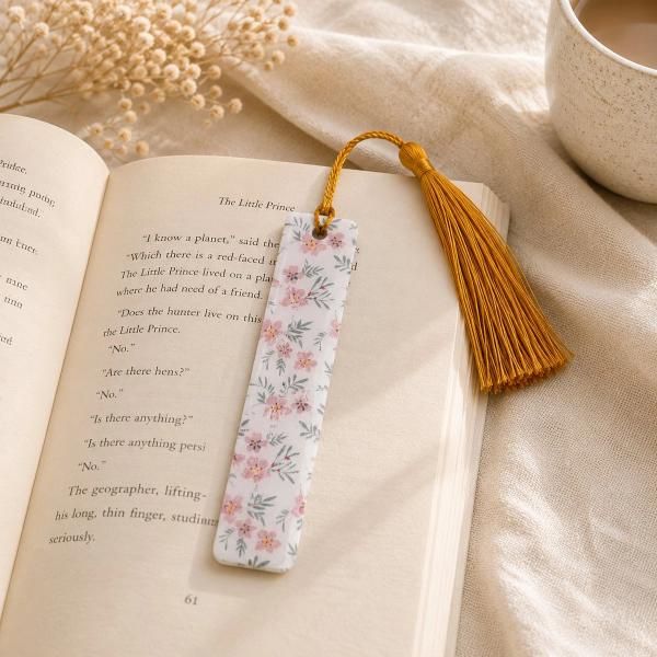 A slim white bookmark printed with pink cherry blossoms and grey leaves, topped with a gold tassel, resting open on a copy of The Little Prince.