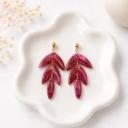 A pair of tiered magenta clay leaf dangle earrings with gold glitter details, displayed on a white ceramic dish with dried flowers in the background.