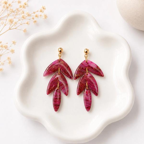 A pair of tiered magenta clay leaf dangle earrings with gold glitter details, displayed on a white ceramic dish with dried flowers in the background.