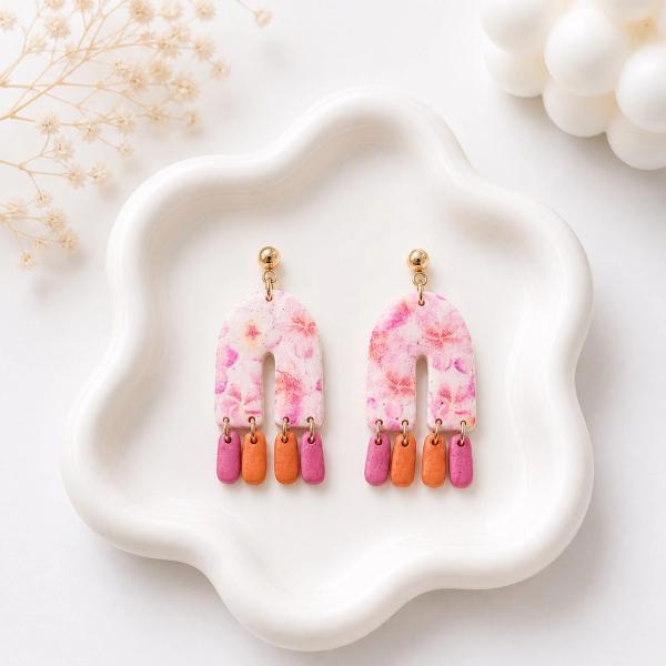 A pair of handmade polymer clay dangle earrings with a white and pink floral arch-shaped top and three small pink and orange rectangular drops hanging from gold-tone rings, displayed on a white ceramic tray.