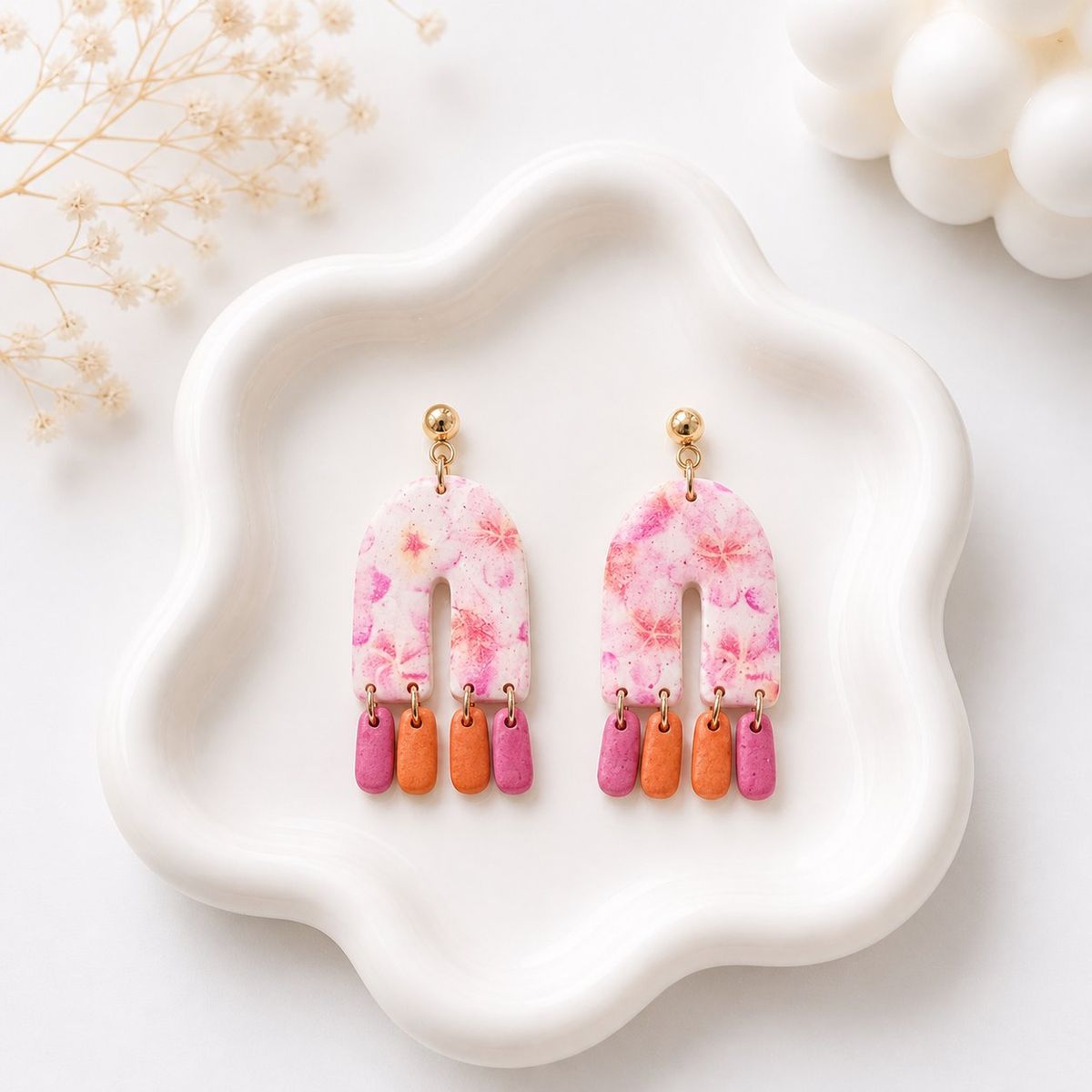 A pair of handmade polymer clay dangle earrings with a white and pink floral arch-shaped top and three small pink and orange rectangular drops hanging from gold-tone rings, displayed on a white ceramic tray.