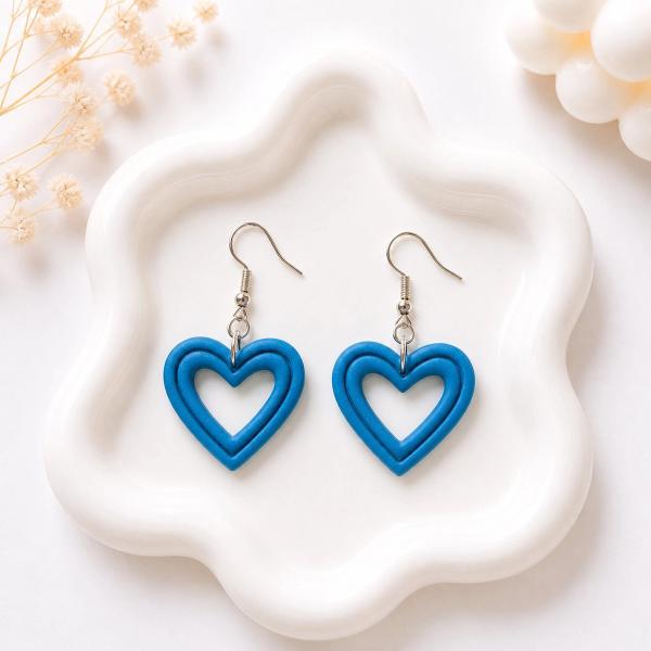 A pair of blue polymer clay open-heart dangle earrings with silver fish hook wires, displayed on a white wavy ceramic tray.