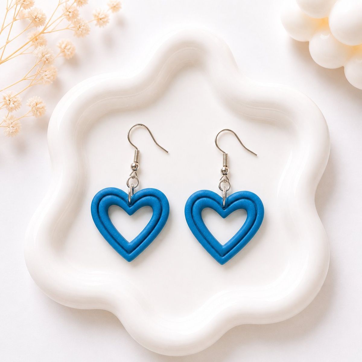 A pair of blue polymer clay open-heart dangle earrings with silver fish hook wires, displayed on a white wavy ceramic tray.