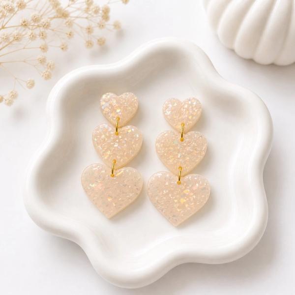 Three graduating cream iridescent glitter heart-shaped clay dangles connected by gold jump rings, displayed on a white scalloped dish.