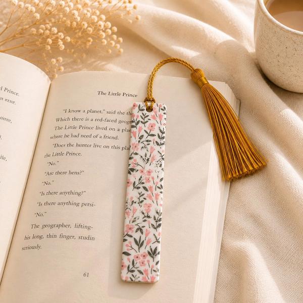 A slim white bookmark printed with pink flowers and dark green botanical stems, topped with a gold tassel, resting on an open book.