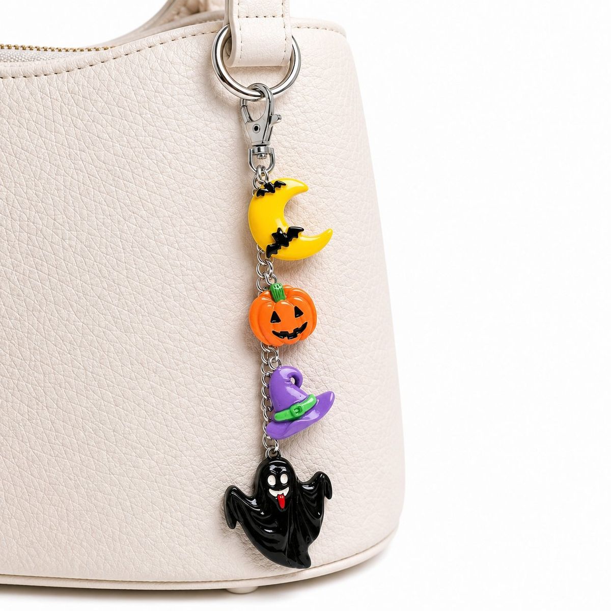 A Halloween-themed bag charm with four resin charms — a yellow crescent moon, orange jack-o'-lantern, purple witch hat, and black ghost — hanging from a silver lobster clasp on a cream handbag.