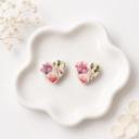 A pair of heart-shaped clay stud earrings with a watercolour floral print in pink, purple, and green, displayed on a white ceramic dish.