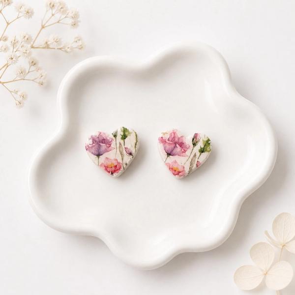 A pair of heart-shaped clay stud earrings with a watercolour floral print in pink, purple, and green, displayed on a white ceramic dish.