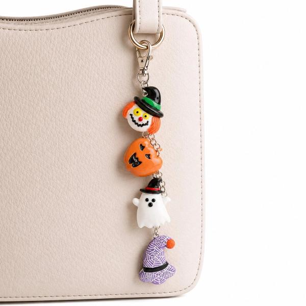 A Halloween-themed keychain charm with four resin figures — a clown with orange hair, an orange jack-o'-lantern, a white ghost wearing a witch hat, and a purple spider-web witch hat — dangling from a silver lobster clasp attached to a cream handbag.