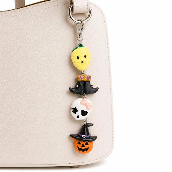 A polymer clay key charm with four Halloween figures — a yellow ghost, black witch boots, a white kawaii skull with a pink bow, and an orange jack-o'-lantern with a black witch hat — clipped to a cream handbag via a silver lobster clasp.