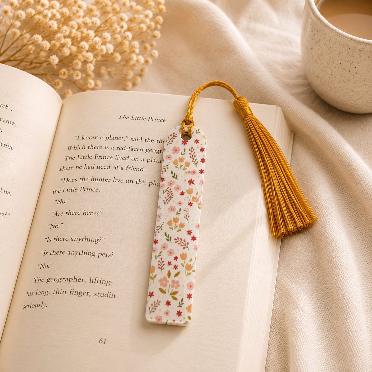 A white rectangular bookmark with a scattered ditsy floral pattern in pink, red, orange, and green, topped with a gold tassel, resting on an open book.