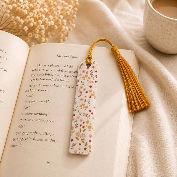 A white rectangular bookmark with a scattered ditsy floral pattern in pink, red, orange, and green, topped with a gold tassel, resting on an open book.