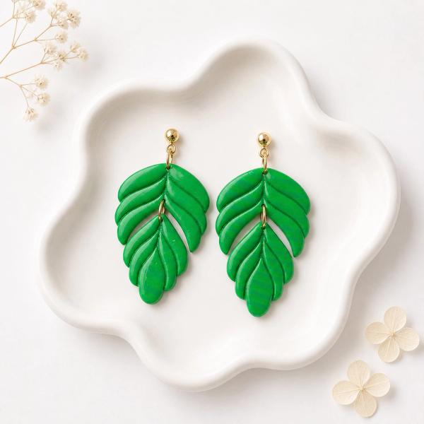A pair of bright green polymer clay leaf dangle earrings with ridged vein detail, attached to gold-tone ball studs, displayed on a white ceramic tray.