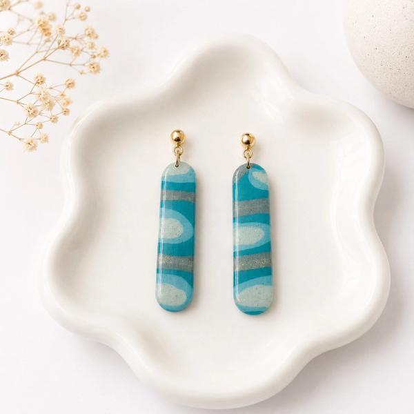 A pair of elongated rounded-bar clay dangle earrings with a teal, light blue, and glittery grey swirl pattern, hanging from gold-tone ball studs, displayed on a white ceramic tray.