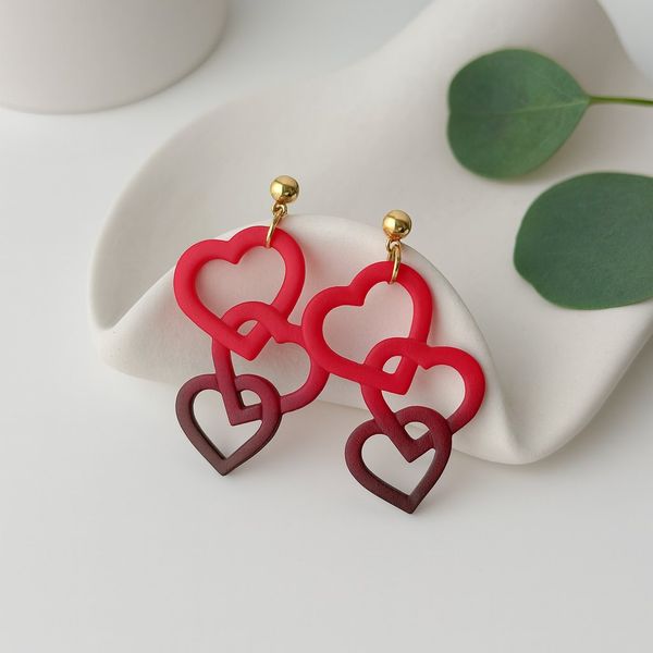 A pair of dangle earrings with two interlocked open heart cutouts in red and dark burgundy, hanging from small gold ball studs, displayed on a white ceramic prop.